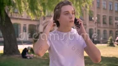 年轻的帅哥穿着白色衬衫打电话，站在大学附近的公园里，<strong>男学生</strong>打电话
